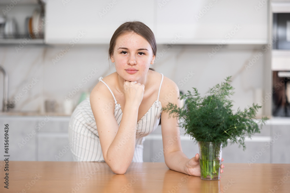 Smiling young girl putting a glass containing bunch of fennel on table in the kitchen with white interior