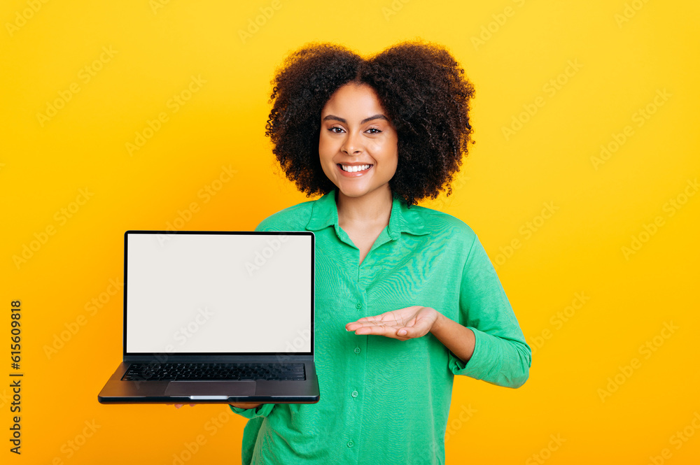 Joyful brazilian or african american curly woman, in green shirt, holding an open laptop in hand with white blank mock up screen and points hand at it, smile at camera, isolated yellow background Joyful brazilian or african american curly woman, in green shirt, holding an open laptop in hand with white blank mock up screen and points hand at it, smile at camera, isolated yellow background