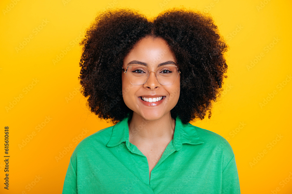 Close-up photo of a pretty, positive curly young woman of brazilian or african american ethnicity, with glasses, in green shirt, looks at camera, smiling friendly, stand on isolated yellow background Close-up photo of a pretty, positive curly young woman of brazilian or african american ethnicity, with glasses, in green shirt, looks at camera, smiling friendly, stand on isolated yellow background