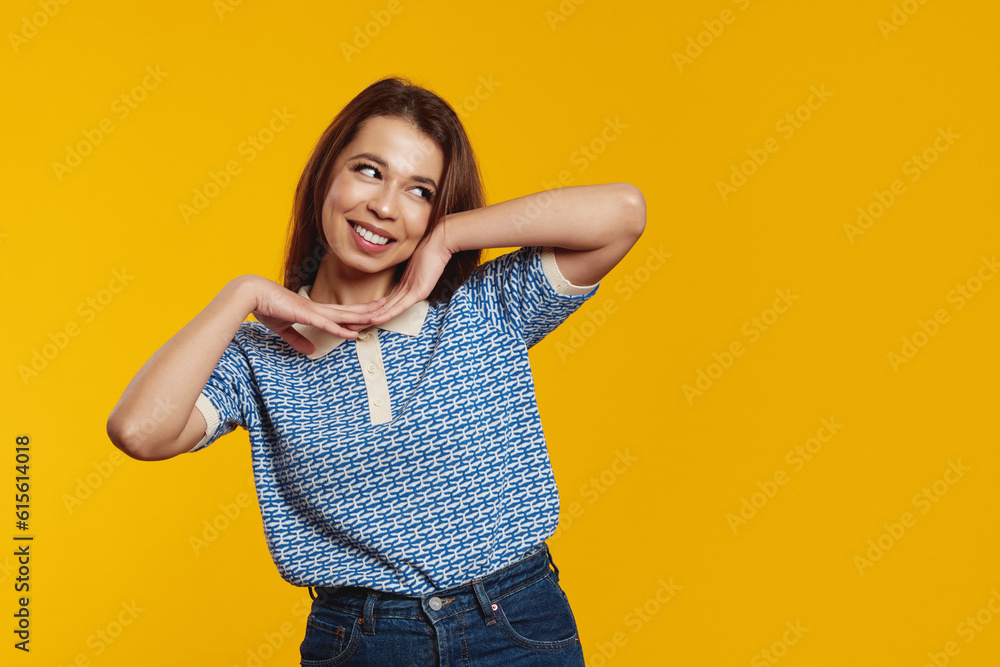 Happy young girl with joyful expression, smiling while holding hands under the chin, looking at free empty space for text against yellow background Happy young girl with joyful expression, smiling while holding hands under the chin, looking at free empty space for text against yellow background