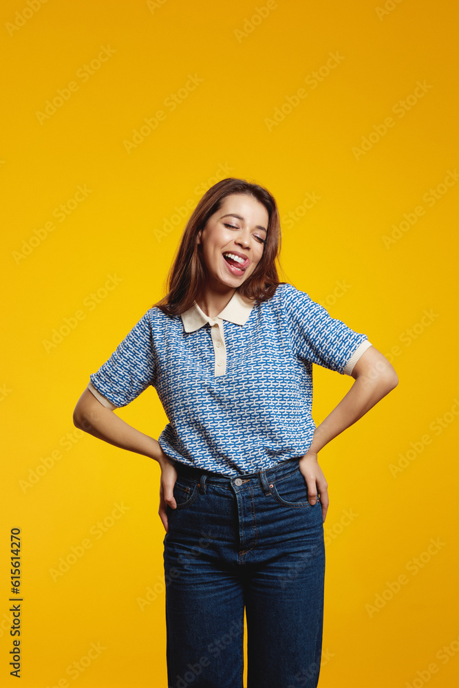 Vertical photo of a happy young girl holding hands on waist while sticking her tongue out isolated over yellow background Vertical photo of a happy young girl holding hands on waist while sticking her tongue out isolated over yellow background