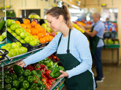 Positive middle-aged saleswoman setting out big peppers on food stall in grocery store