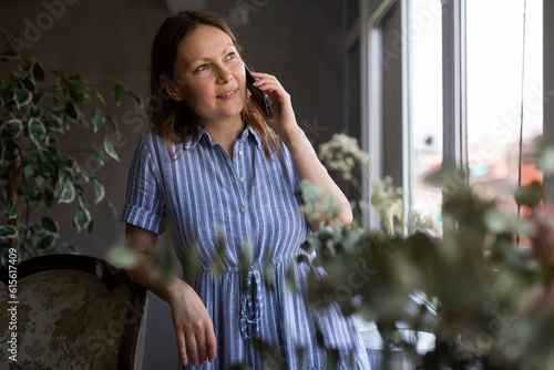 Foto Smiling positive adult woman talking on mobile phone at the window in her house