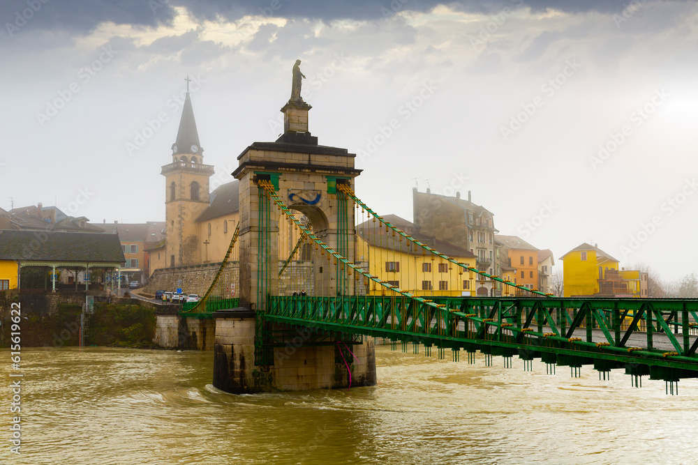 View of Seyssel townscape on bank of Rhone river with ancient Saint-Blaise church and suspension bridge on winter day, Haute-Savoie department, France