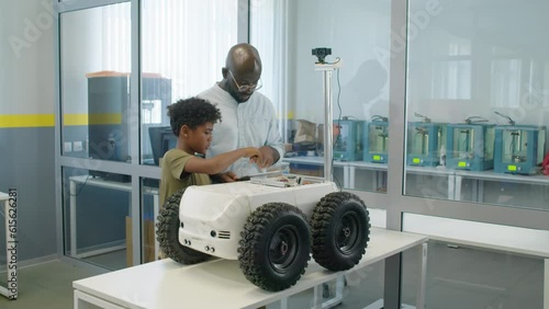 Full shot of African American boy and man in glasses standing next to delivery robot in lab, looking at its circuitry and wires, checking with diagram on tablet computer, and discussing its design