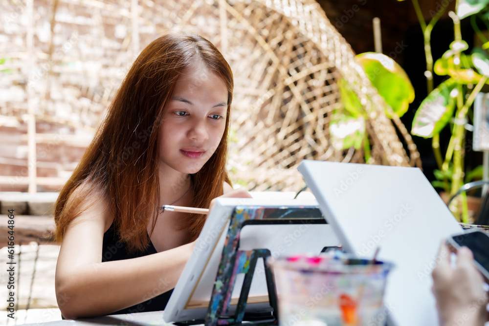 Portrait of beautiful Asian painter with long hair intending to paint sitting at table holding paintbrush In art class to practice your skills on a table with paint and a white canvas for painting. Portrait of beautiful Asian painter with long hair intending to paint sitting at table holding paintbrush In art class to practice your skills on a table with paint and a white canvas for painting.