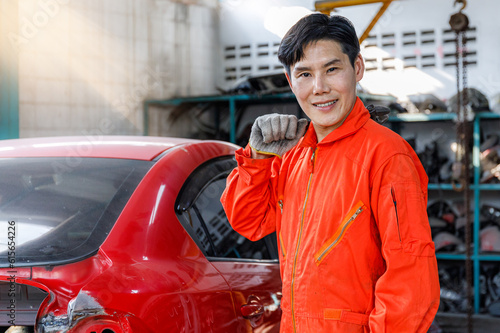 Young Asian worker in orange uniform looking at camera smiling. Skilled worker holding wrench on his shoulder working in auto workshop.
