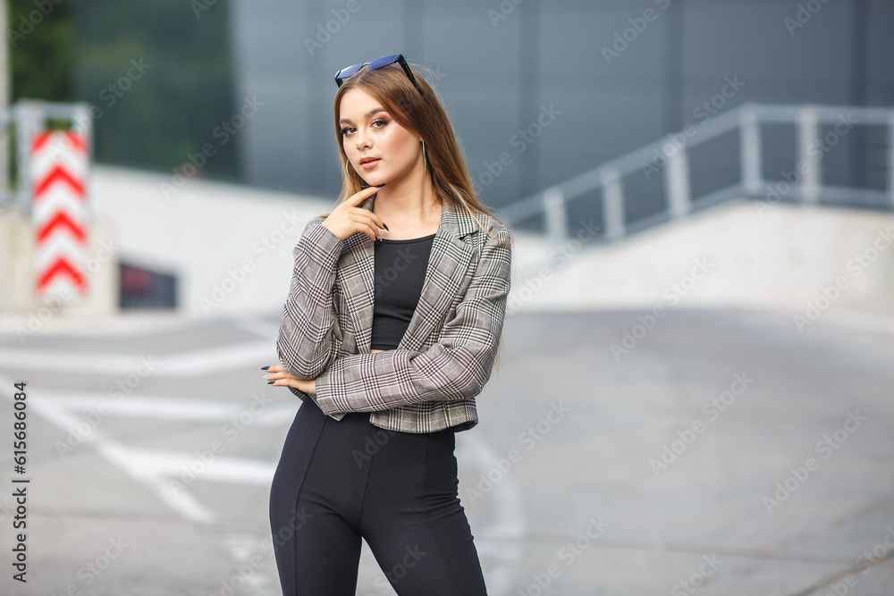 Fototapeta premium beautiful young girl with sunglasses in jacket posing near a modern office building with mirrored walls in a parking lot with road signs