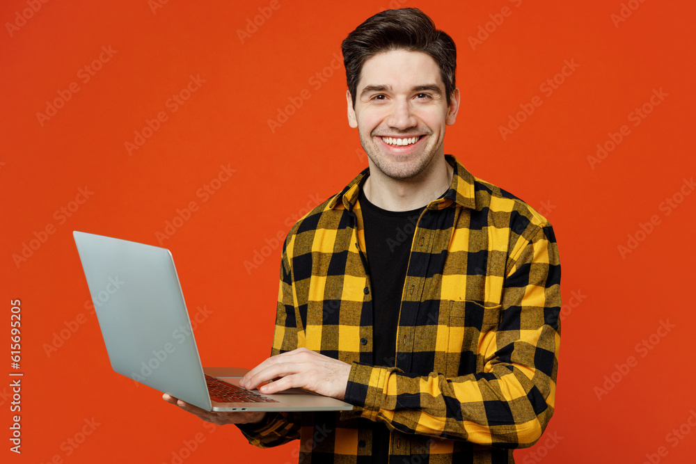 Young smiling happy fun caucasian IT man he wears yellow checkered shirt black t-shirt hold use work on laptop pc computer isolated on plain red orange background studio portrait. Lifestyle concept. Young smiling happy fun caucasian IT man he wears yellow checkered shirt black t-shirt hold use work on laptop pc computer isolated on plain red orange background studio portrait. Lifestyle concept.