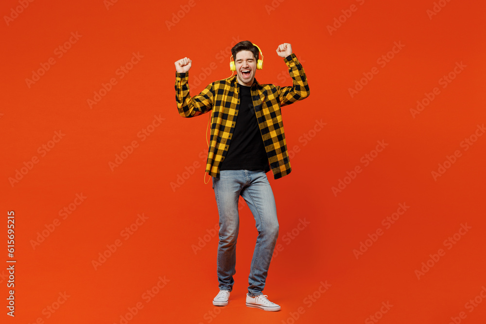 Fototapeta premium Full body young cheerful happy man wear yellow checkered shirt black t-shirt headphones listen to music raise up hands dance isolated on plain red orange background studio portrait. Lifestyle concept.