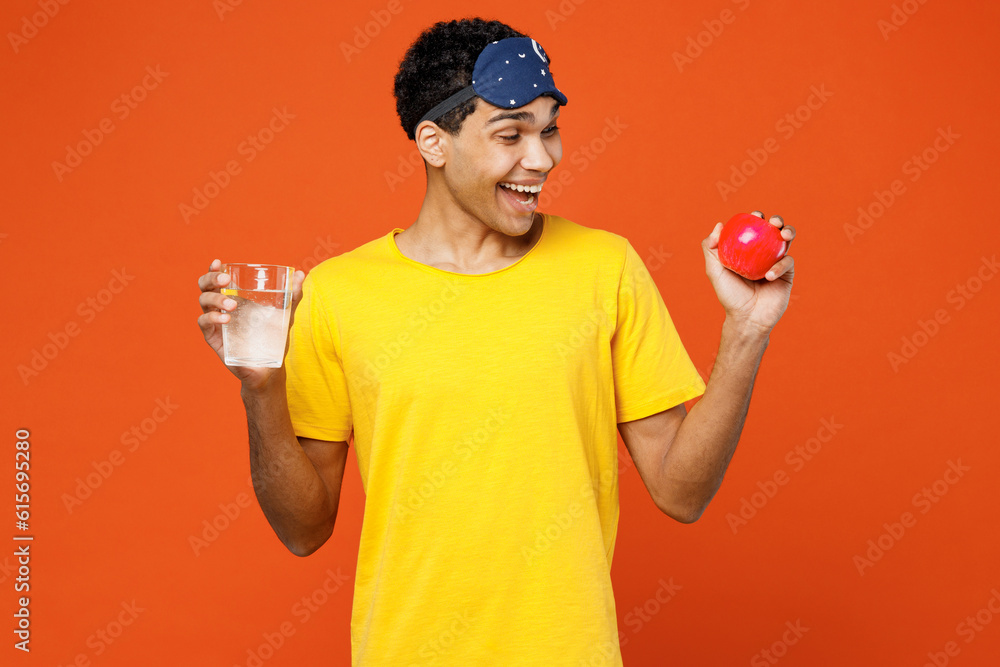 Calm surprised fun excited young man wear pyjamas jam sleep eye mask rest relax at home hold in hand glass of water red apple isolated on plain orange background studio. Good mood night nap concept. Calm surprised fun excited young man wear pyjamas jam sleep eye mask rest relax at home hold in hand glass of water red apple isolated on plain orange background studio. Good mood night nap concept.