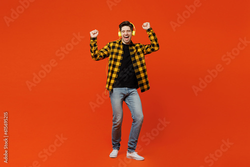 Full body young cheerful happy man wear yellow checkered shirt black t-shirt headphones listen to music raise up hands dance isolated on plain red orange background studio portrait. Lifestyle concept.