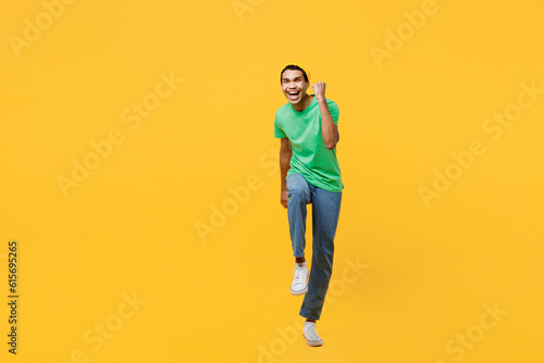 Full body young man of African American ethnicity he wears casual clothes green t-shirt hat doing winner gesture celebrate clenching fists say yes isolated on plain yellow background studio portrait.