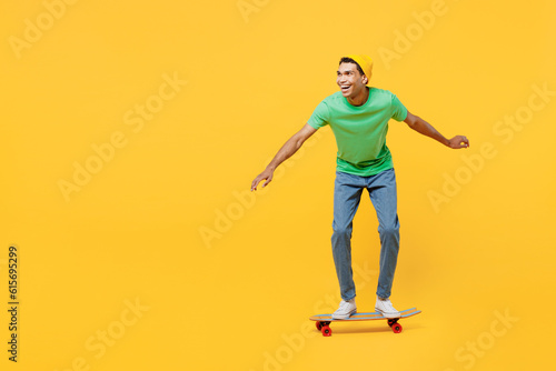 Full body side view smiling young man of African American ethnicity he wears casual clothes green t-shirt hat rising skateboard isolated on plain yellow background studio portrait. Lifestyle concept.