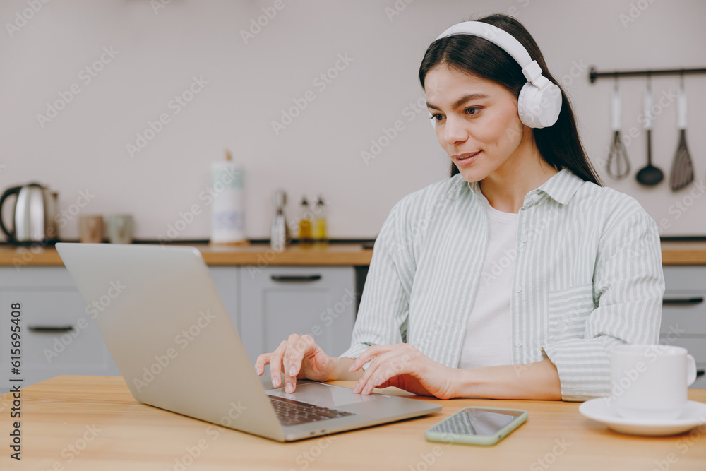 Young happy housewife latin IT woman wear casual clothes headphones shirt hold use work on laptop pc computer listen music sit at table in light kitchen at home alone. Lifestyle cooking food concept. Young happy housewife latin IT woman wear casual clothes headphones shirt hold use work on laptop pc computer listen music sit at table in light kitchen at home alone. Lifestyle cooking food concept.