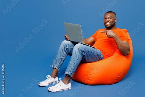 Full body young IT man of African American ethnicity he wear orange t-shirt sit in bag chair hold use work point on laptop pc computer isolated on plain dark royal blue background. Lifestyle concept.