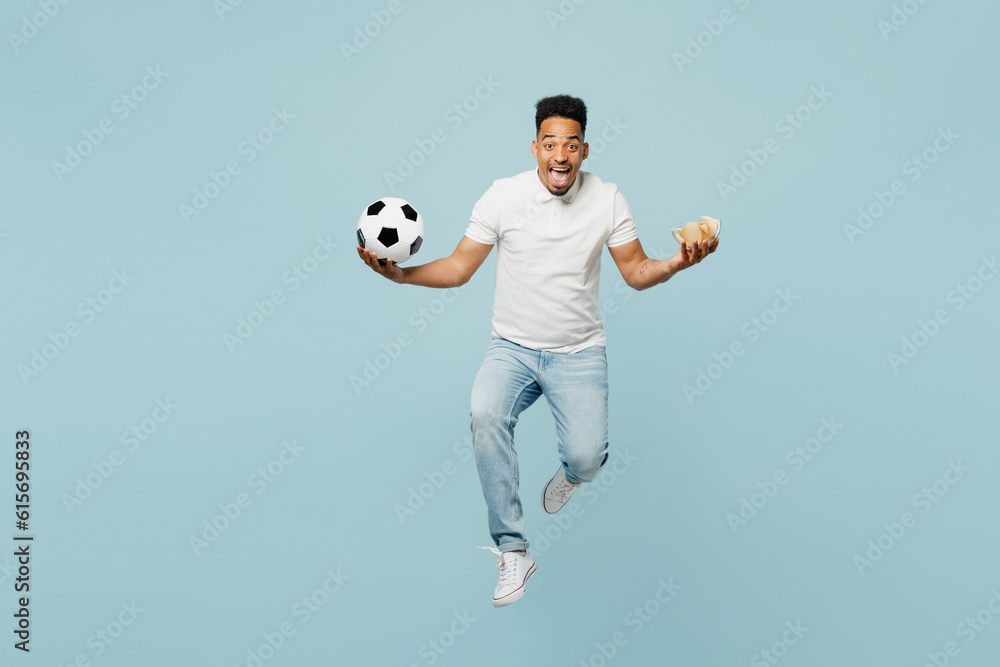 Full body excited young man fan wear t-shirt cheer up support football sport team hold in hand chips in bowl soccer ball jump high watch tv live stream isolated on plain pastel blue color background.