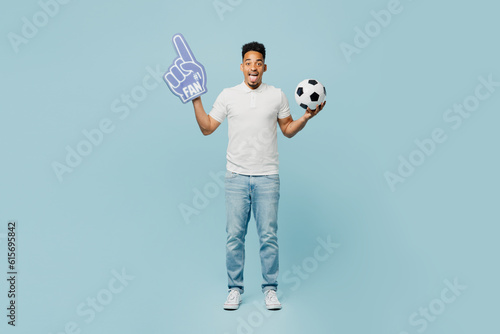 Full body young man fan wear basic t-shirt foam 1 fan glove finger up cheer up support football sport team hold in hand soccer ball watch tv live stream isolated on plain pastel blue color background. photo
