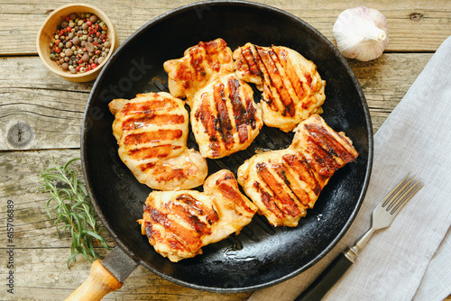 View from above. Grilled chicken thigh fillet with strips on a rustic wooden table in a cast iron pan, with rosemary, garlic pepper and fork. Flat lay.