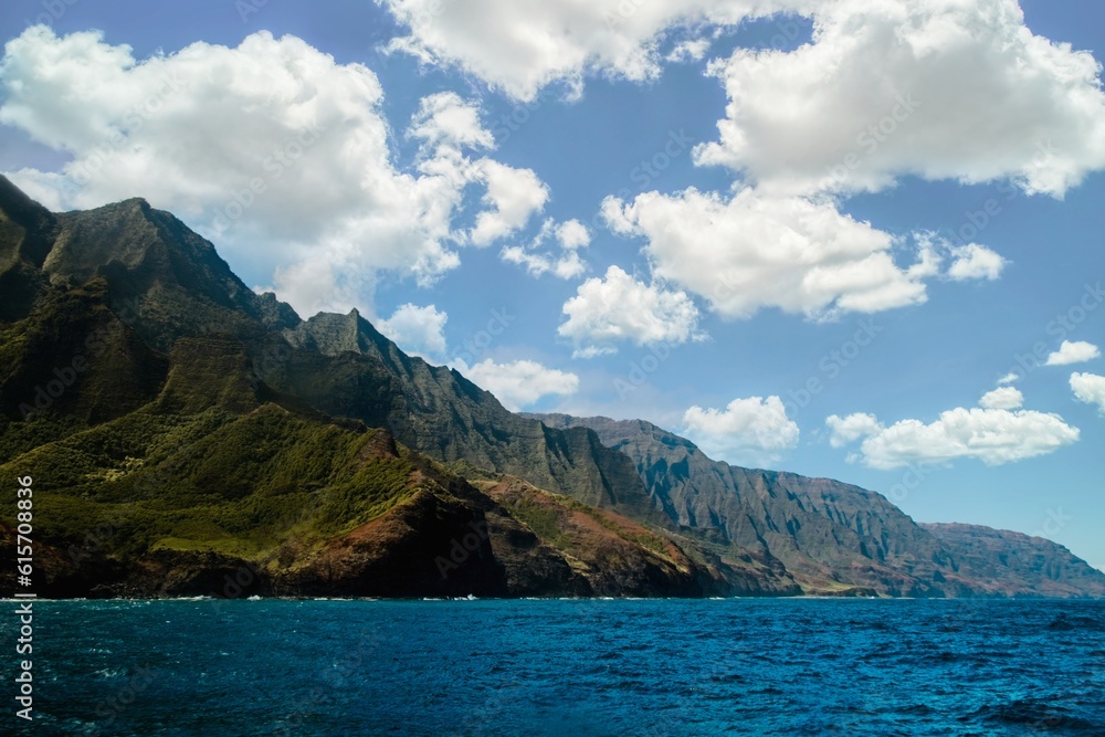 View of Na Pali coast of the island of Kauai in Hawaii from the sea with mountains and ocean as background and marshmallow clouds.