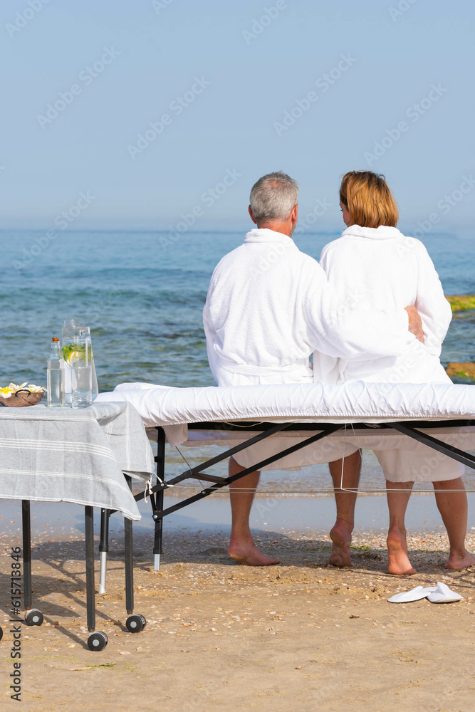 Obraz premium Family – man and woman sitting on massage table and looking at the sea after having relax procedures in tropical massage spa on the beach.