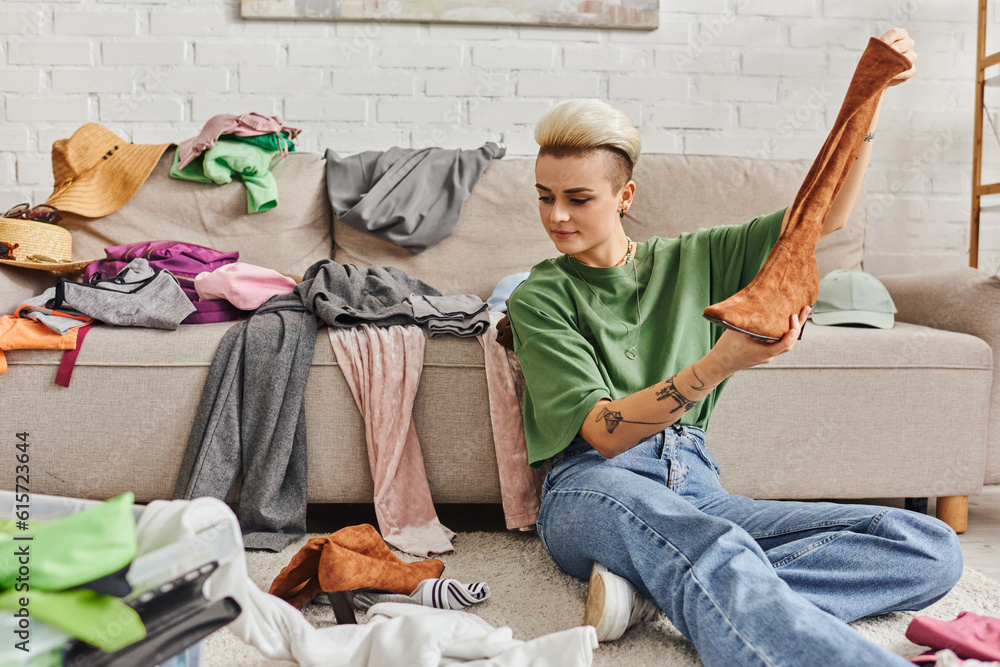 Obraz premium young woman sitting on floor with suede boot near clothes and couch at home, sorting and reducing wardrobe, trendy hairstyle, tattoo, sustainable living and mindful consumerism concept