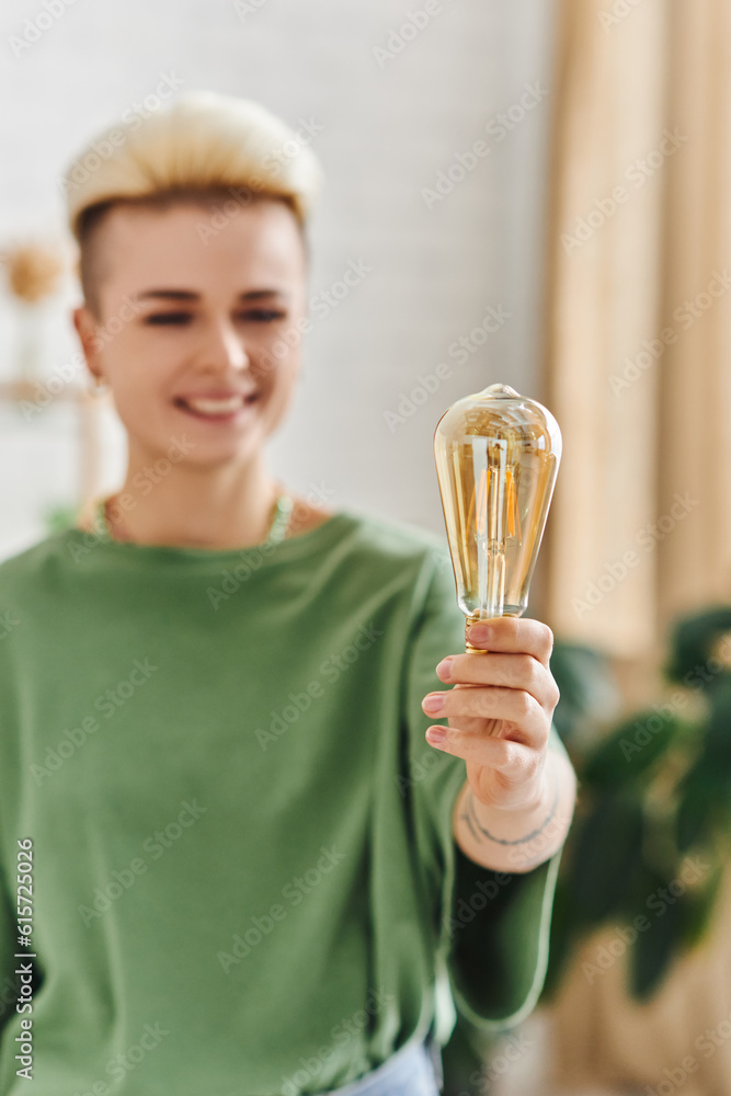 cheerful woman with trendy hairstyle, in casual clothes smiling and standing with energy saving light bulb at home on blurred background, sustainable lifestyle and environmentally conscious concept cheerful woman with trendy hairstyle, in casual clothes smiling and standing with energy saving light bulb at home on blurred background, sustainable lifestyle and environmentally conscious concept