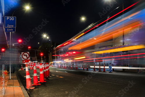 Orange traffic cones lined up on the road. Bus light trails approaching red traffic lights. Auckland.