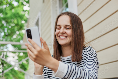 Cheerful caucasian attractive adult woman wearing striped shirt using phone sitting on porch of house reading comments in social networks looking at device display.