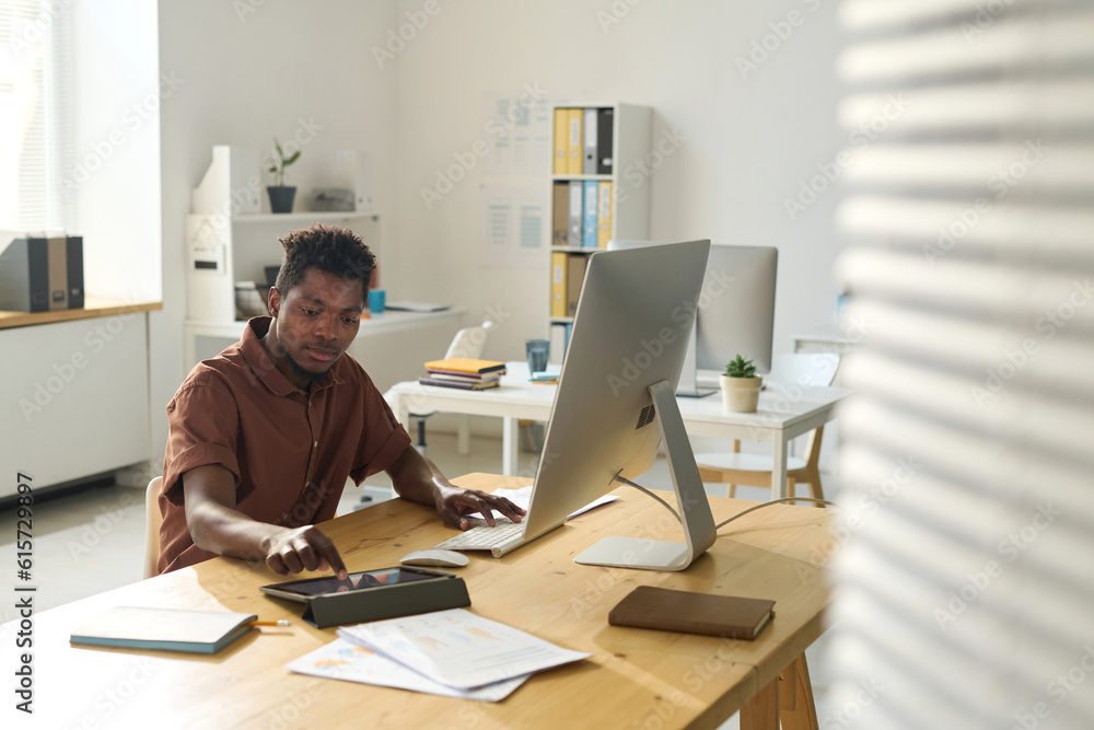 African American businessman working on tablet pc while sitting at his workplace with computer monitor in office