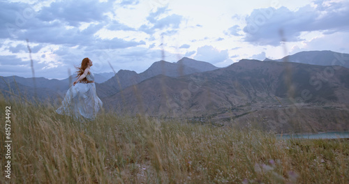 Young beautiful girl with red hair wearing white dress walking on top of a mountain facing wind blowing her hair and dress - freedom, adventure, harmony. Copy space