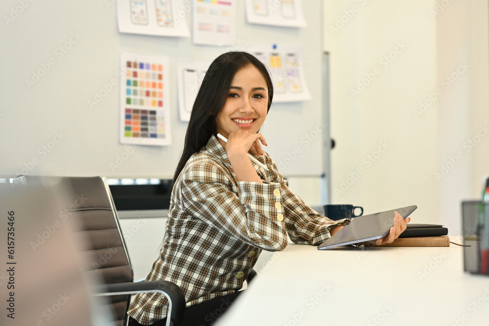 Portrait of confident female web developer holding digital tablet, sitting at meeting table in creative office and smiling to camera Portrait of confident female web developer holding digital tablet, sitting at meeting table in creative office and smiling to camera