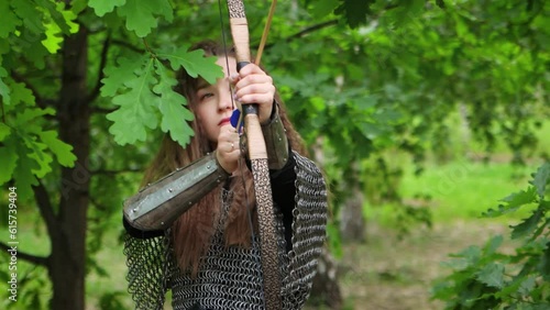 Smooth camera movement, selective focus, portrait of a young woman in chain mail and bracers drawing a bowstring and aiming a bow, against the backdrop of a summer forest.