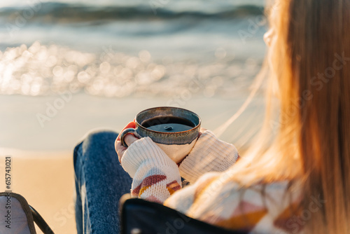 Closeup back portrait of a woman sitting in chair on winter sea shore in a cozy sweater and holding hot cup of coffee. A girl enjoying beautiful view of autumn ocean seaside waterfront waves.