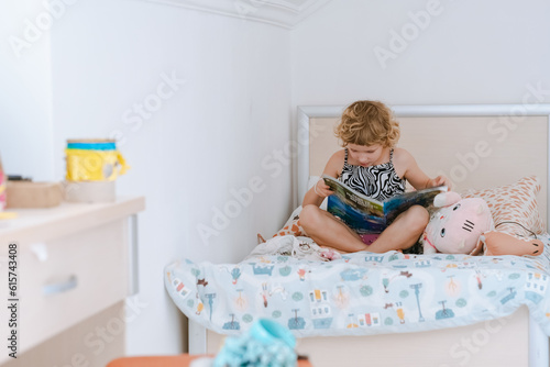 Cute little girl reading by herself an educational magazine on the bed in the bedroom.