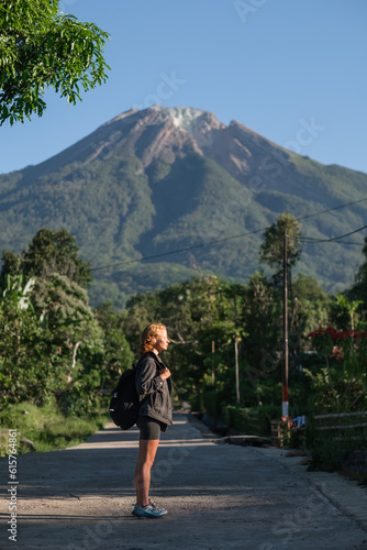 A beautiful, young girl, of European appearance, with blond, curly hair, is traveling with a backpack hitchhiking around the island overlooking the volcano.