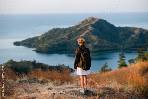 A young girl, blonde, with a backpack, stands on top of a mountain, rear view, overlooking a beautiful panorama of the islands and bays in Indonesia. © Evgenii