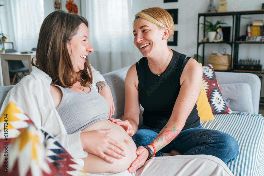 Young woman tender touching partner's female pregnant belly. Same-sex marriage couple on home living room sofa. Woman's health, happy pregnancy doula supporting and calm mental mood concept image Young woman tender touching partner's female pregnant belly. Same-sex marriage couple on home living room sofa. Woman's health, happy pregnancy doula supporting and calm mental mood concept image