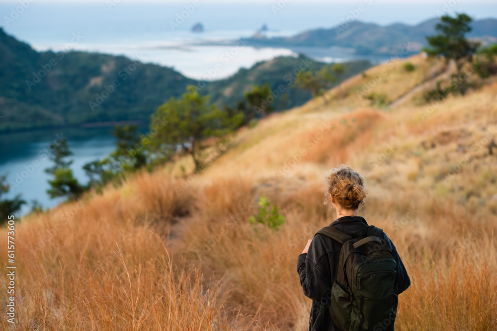 A young girl, blonde, with a backpack, stands on top of a mountain, rear view, overlooking a beautiful panorama of the islands and bays in Indonesia.