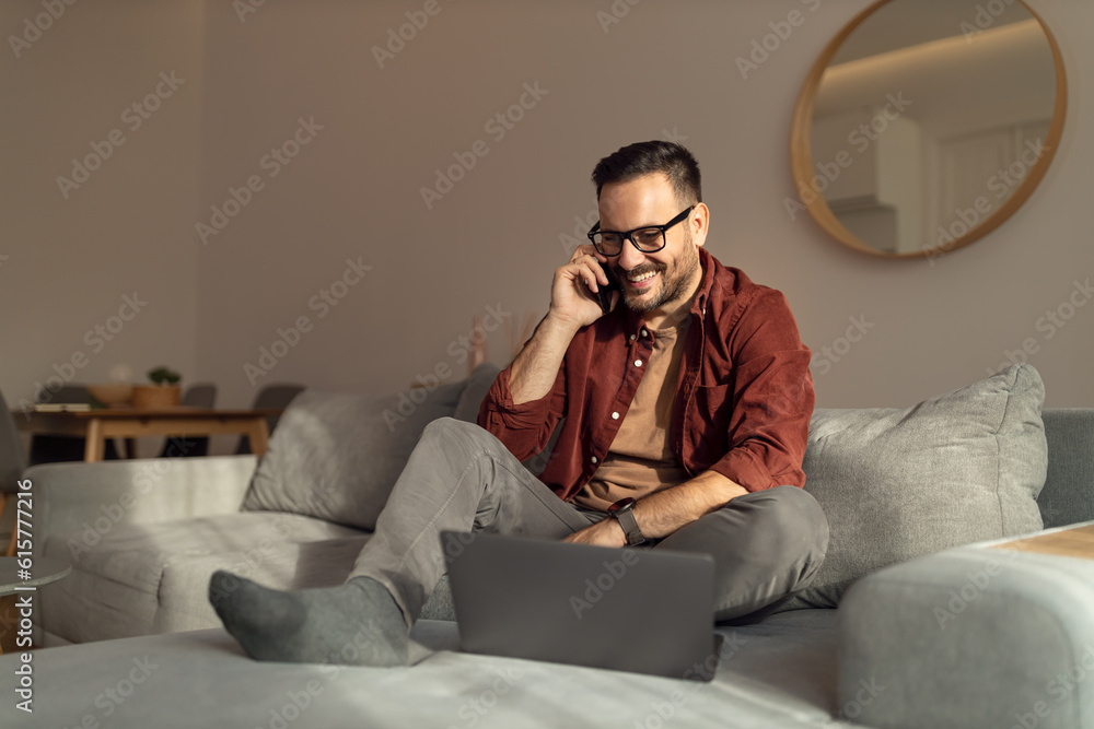 Smiling man working over the laptop while making a phone call, sitting on the couch at home.