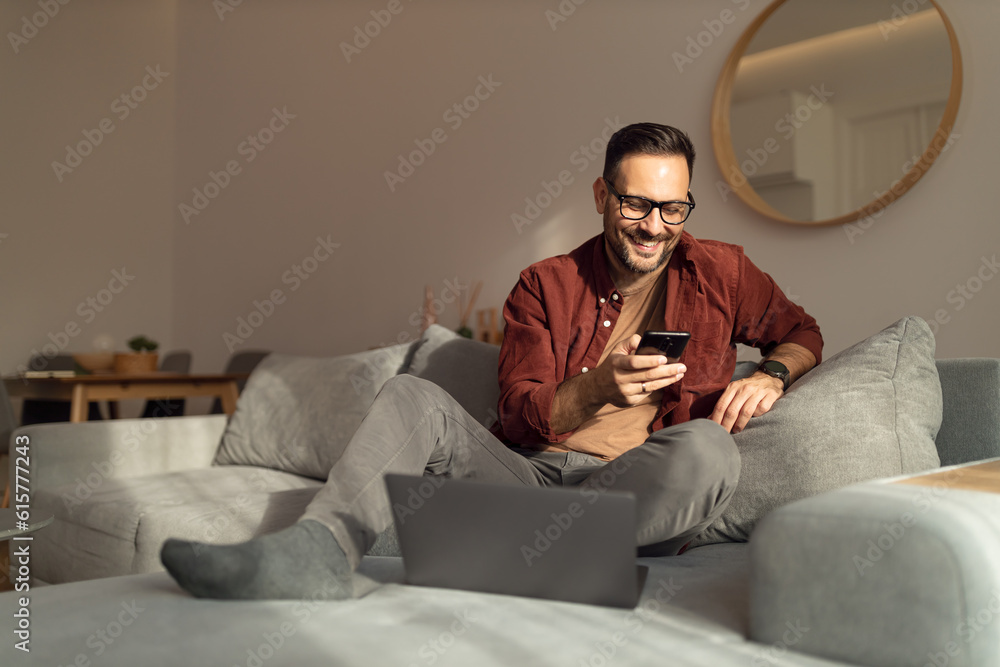 Smiling man using a mobile phone, sitting on sofa, being at home on a sunny day.