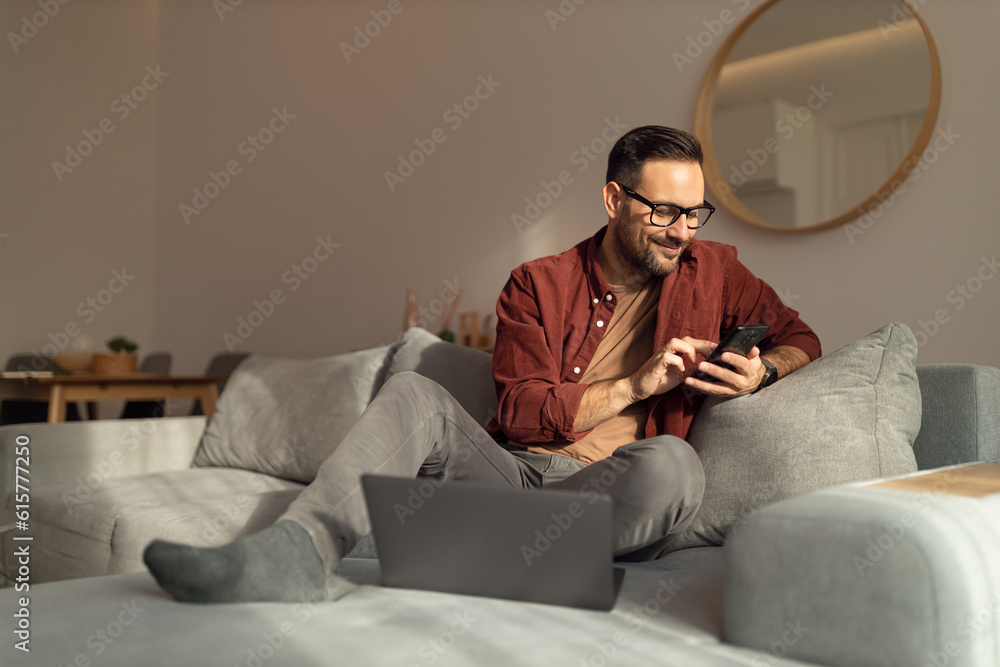 Obraz premium Smiling man checking his social medias, wearing glasses, dressed casually, sitting on the sofa.