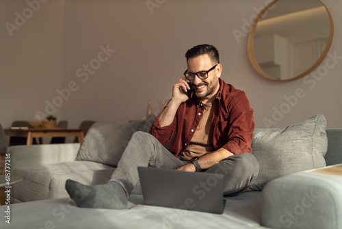 Smiling man working over the laptop while making a phone call, sitting on the couch at home.