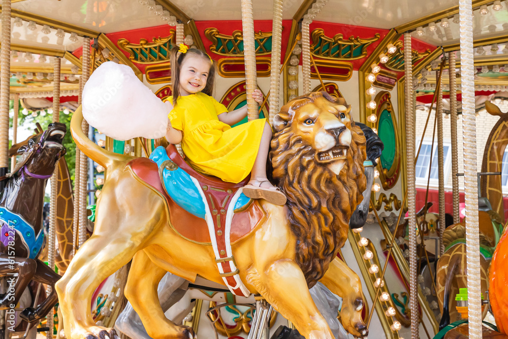 Adorable little girl in summer yellow dress at amusement park having a ride on the merry-go-round and eating cotton candy