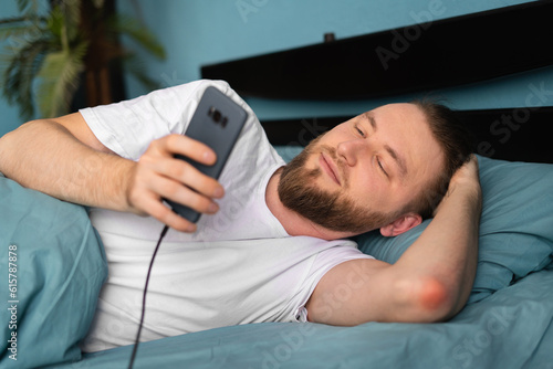 Caucasian man texting on cellphone and browsing internet lying in ded in bedroom indoors. Guy uses mobile app via phone. Technology and gadgets photo