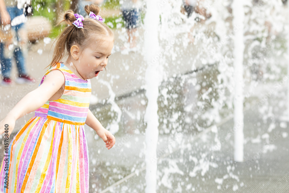 Naklejka premium A happy little girl has fun near the city fountain. The child has a rest near a fountain during a heat wave.