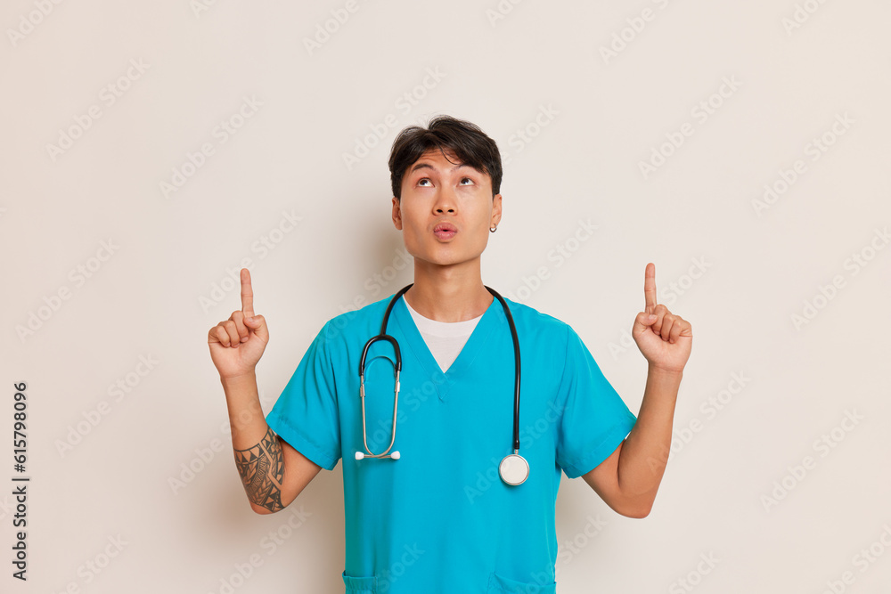 Young asian male doctor in medical uniform poses on white background, looks up and points with index fingers up, professional people concept, copy space Young asian male doctor in medical uniform poses on white background, looks up and points with index fingers up, professional people concept, copy space