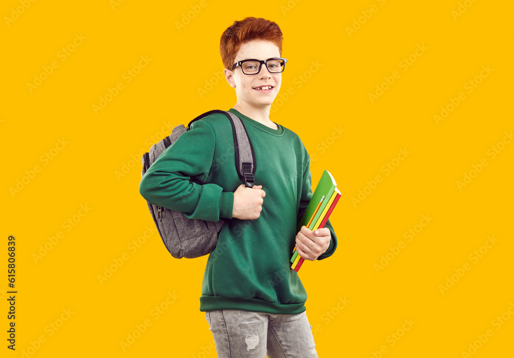 Smiling handsome teenage boy with backpack holding books. Positive redhead schoolboy wearing glasses, green sweatshirt and jeans posing over isolated yellow background. School education concept Smiling handsome teenage boy with backpack holding books. Positive redhead schoolboy wearing glasses, green sweatshirt and jeans posing over isolated yellow background. School education concept