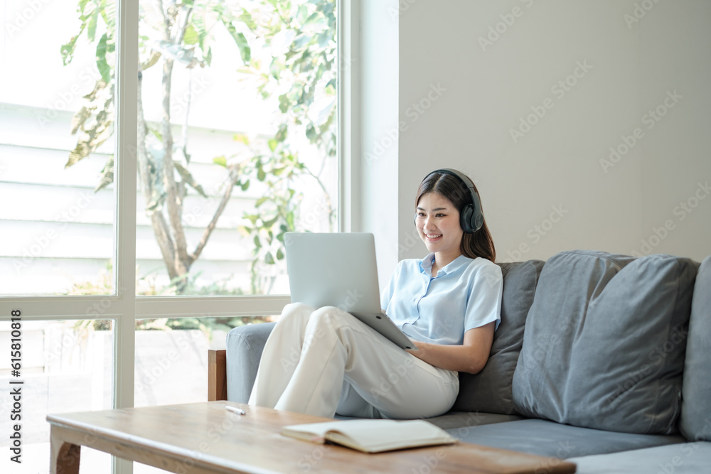 Beautiful Young asian woman at home sitting on the sofa using laptop and headphone listening to music at home.