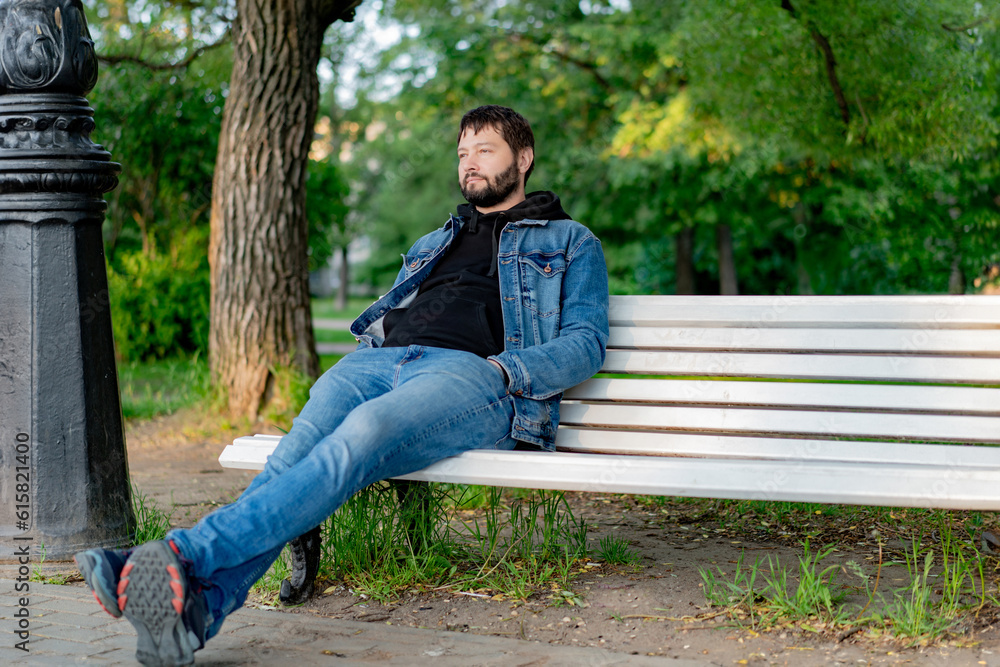 attractive caucasuan bearded young man sitting on bench in park looking aside